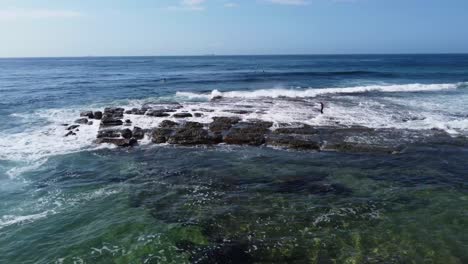Aerial-drone-landscape-shot-of-kid-surfer-walking-over-rocks-reef-Central-Coast-waves-Pacific-Ocean-NSW-Australia-4K