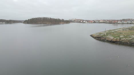 Flying-over-the-still-water-in-Karlskrona,-Sweden-where-you-can-see-the-beautiful-cityscape-in-the-background-and-then-the-amazing-picturesque-little-island-of-Brandaholm-with-the-small-red-cabins