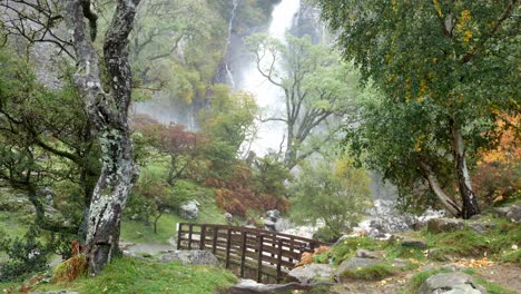 Idyllic-wooden-bridge-over-valley-waterfall-cascading-into-powerful-flowing-river-wide-shot