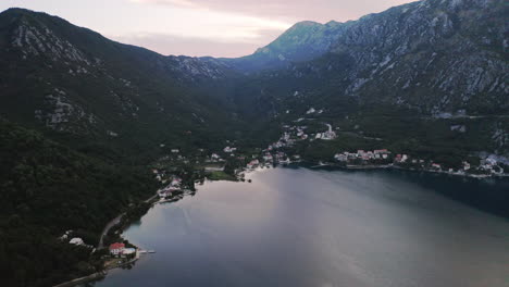 A-coastal-town-on-the-shores-of-the-bay-of-Kotor-in-Montenegro,-surrounded-by-high-mountains-covered-with-vegetation,light-morning-glow-above-the-peaks,-buildings-and-trees-on-the-mountainside,aerial