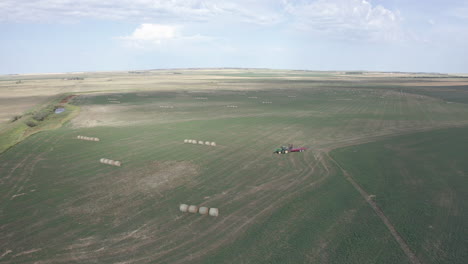Sensational-scenic-flight-above-vast-open-green-farmland-towards-industrial-farming-machine-hauling-bales-of-hay-in-flat-expansive-rural-countryside,-Saskatchewan,-Canada,-overhead-aerial-approach