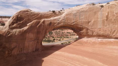 Aerial-backward-of-man-walking-over-Wilson-Arch-rock-formation,-Utah
