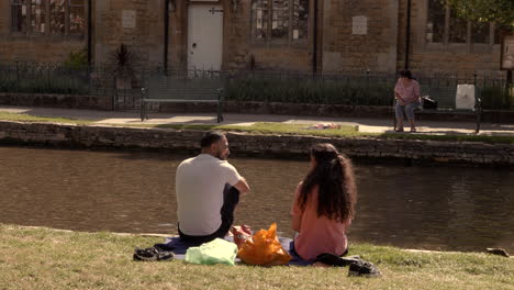 During-the-COVID-19-pandemic,-a-couple-with-others-is-seen-enjoying-a-sunny-day-by-the-side-of-a-canal-in-Bourton-on-The-Water,-Cotswold,-United-Kingdom,-while-maintaining-social-distancing