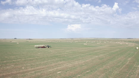 Vuelo-Panorámico-Sobre-Tierras-De-Cultivo-Verdes-Por-Parte-De-Un-Agricultor-Que-Conduce-Una-Máquina-De-Tractor-Agrícola-Industrial-Que-Transporta-Fardos-De-Heno-En-Un-Campo-Rural-Plano-Y-Expansivo-Y-Un-Gran-Cielo,-Saskatchewan,-Canadá,-Círculo-Aéreo