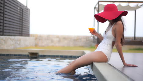 Frau-Mit-Rotem-Hut,-Die-Sich-Neben-Dem-Pool-Entspannt-Und-Einen-Orangencocktail-Trinkt