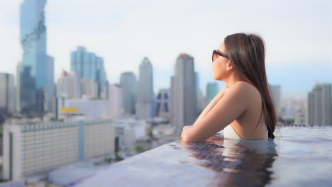 While-leaning-on-the-edge-of-a-rooftop-resort-infinity-pool-an-attractive-woman-looks-out-on-the-city-skyline-of-Bangkok,-Thailand