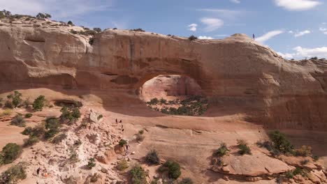Group-Of-Travelers-Climbing-The-Amazing-Red-Rock-Formation-Known-As-Wilson-Arch,-A-Roadside-Attraction-In-Moab,-Utah,-United-States---Wide-Shot-Pan-Left