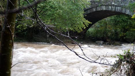 Inundaciones-Rápidas-Que-Fluye-El-Agua-Del-Río-Bajo-El-Idílico-Puente-De-Piedra-Arqueado-Rural-Dolly-A-La-Derecha