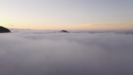 Drone-flies-over-a-layer-of-clouds-where-the-top-of-a-mountain-protrudes-during-a-sunrise