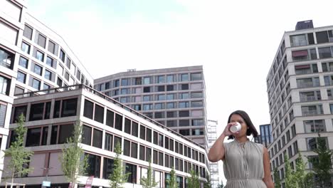 Young-business-woman-drinks-coffee-to-go-while-walking-between-tall-office-buildings-in-financial-district-during-her-break