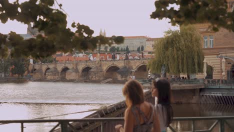Two-tourist-girls-talking,-Charles-Bridge-in-background,-sunny-day-in-Prague,-slow-motion