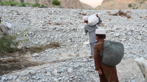 Video-Von-Zwei-Kleinen-Kindern-Und-Einem-Mann,-Die-Schwere-Säcke-Auf-Ihren-Schultern-Tragen-Und-Auf-Der-Kaputten-Straße-In-Baloschistan-Gehen