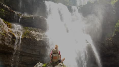 Girl-hiking-is-sitting-on-a-rock-under-a-waterfall-in-a-nature-reserve