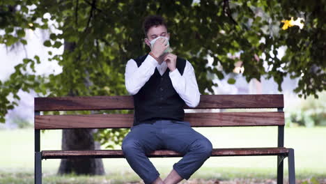 An-elegant-young-man-in-a-park-wearing-a-black-waistcoat-and-white-shirt-putting-on-a-green-protective-Covid-19-mask,-sitting-on-a-park-bench-and-casually-relaxing-before-standing-up,-static-4k-shot