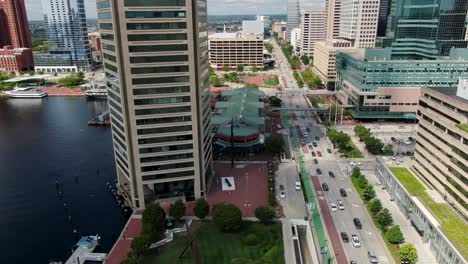 Rising-aerial-features-Top-of-the-World-Observation-building,-Inner-Harbor,-Pratt-Street-traffic-on-summer-day