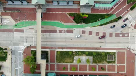 Top-down-overhead-aerial-drone-view,-cars-on-street,-red-brick-sidewalk,-catwalk-pedestrian-bridge-across-road,-traffic-drives-through-intersection