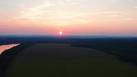 Beautiful-pink-orange-sun-peeks-through-the-clouds-and-reflects-of-amazing-Potomac-River-in-Maryland-surrounded-by-farms,-Drone-Aerial