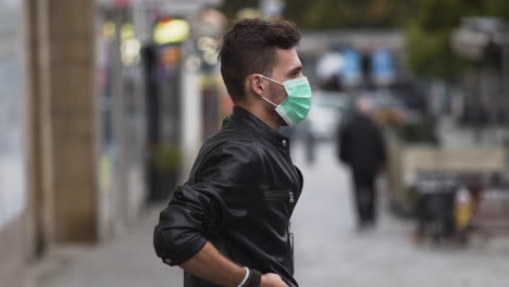 A-young-man-in-a-leatherette-jacket-with-a-modern-hairstyle-on-a-busy-street,-putting-on-a-green-protective-Covid-19-facemask,-putting-his-hands-in-his-pockets,-sideways-to-the-camera,-close-up-4k