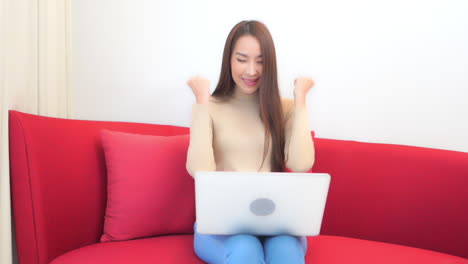 Asian-lady-sitting-with-a-wireless-laptop-on-her-knees-on-a-red-sofa-in-her-living-room-using-the-device-and-tightening-her-fists-with-joy
