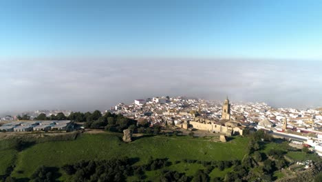 Aerial-dolly-shot-over-the-landscape-in-front-of-medina-sidonia-in-andalucia-spain-with-a-view-of-the-church-of-santa-maria-and-the-beautiful-old-town-with-clouds-and-blue-sky