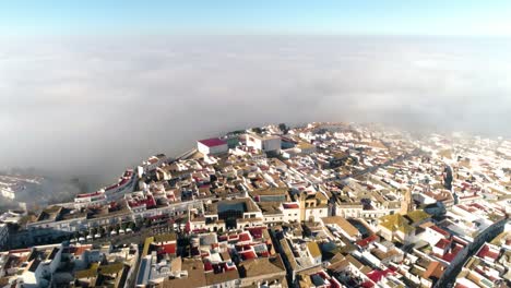 Wide-aerial-drone-shot-over-the-historic-old-town-of-medina-sidonia-in-andalucia-spain-with-predominantly-white-buildings-and-red-roofs-overlooking-the-clouds-and-blue-sky