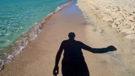 Silhouette-Schatten-Eines-Mannes,-Der-Eine-Sonnenbrille-Aufsetzt-Und-Am-Sandstrand-Entlang-Der-Türkisfarbenen-Meerwasserküste-Spazieren-Geht
