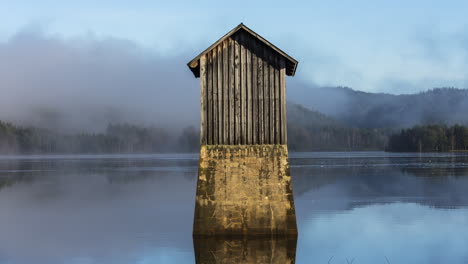 Tiny-old-wooden-cabin-in-middle-of-tranquil-lake-covered-in-thick-fog