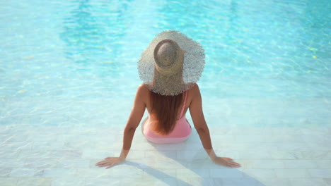 With-her-back-to-the-camera,-a-healthy,-fit-woman,-in-a-straw-sun-hat-and-pink-bathing-suit,-relaxes-in-the-shallow-end-of-a-resort-pool