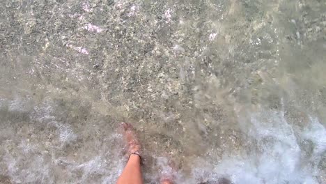 Slow-motion-view-of-two-feet-standing-on-a-sandy-tropical-beach-while-waves-of-clear-warm-water-splash-onto-the-shore