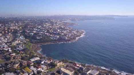 Luftflug-über-Küstenstrand-Küstengrundstück-Ozeanhäuser-Mit-Blick-Auf-Die-Stadt-Im-Hintergrundhorizont-In-Sydney,-Australien