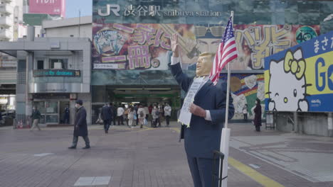 Mann,-Der-Maske-Trägt-Und-Als-Donald-Trump-Verkleidet-Ist,-Steht-Während-Einer-Pandemie-In-Tokio,-Japan,-Auf-Dem-Hachiko-platz-In-Der-Nähe-Des-Bahnhofs-Shibuya-Mit-Passanten