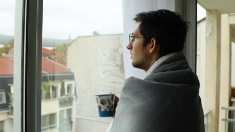 Young-man-resting-at-home-and-looking-through-window-during-overcast-day-with-cosy-blanket-and-cup