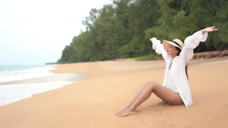 Girl-dressed-in-white-sitting-on-beach-raises-her-arms-smiling-satisfied