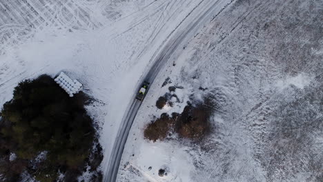 Drone-panning-slowly-above-green-Claas-tractor-driving-slowly-on-slippery-icy-road-across-fields-filled-with-frosty-snow-in-winter-Scandinavian-Nordic-northern-landscape-cold-frozen-morning-near-lake
