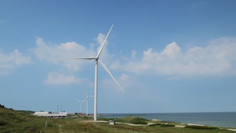 Turbina-Eólica-En-La-Costa-Girando-Lentamente-Con-Mar-Y-Nubes-En-Segundo-Plano