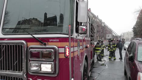 Feuerwehrauto-Mit-Blinkenden-Lichtern-Stationär-Auf-Der-Brooklyn-Street-Unter-Starkem-Schneefall-Mit-Einer-Gruppe-Von-Feuerwehrleuten---Mittlerer-Schuss