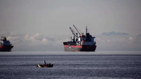 Enormous-empty-bulk-carrier-with-deck-cranes-on-the-anchorage-waiting-for-orders-at-the-background-while-people-in-a-kayak-enjoy-life-on-a-cloudy-day