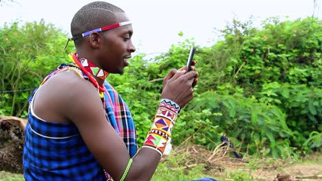 Young-Moran-male-texting-with-a-phone,-sitting-outdoors-in-forests-of-Africa