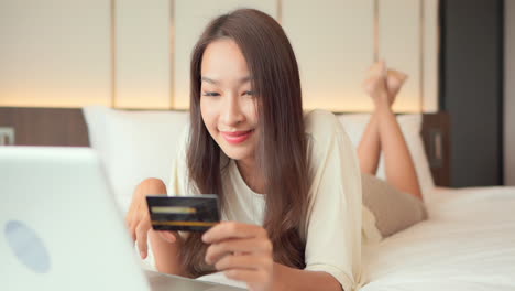 Attractive-Asian-woman-lying-on-king-size-bed-and-using-her-credit-card-for-shopping-online,-type-in-card-information-on-the-mobile-phone-and-winning-prize-lottery,-exited-face-expression,-close-up