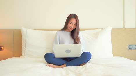 Lächelnde-Asiatische-Frau,-Die-Mit-Laptop-Auf-Bett-Sitzt