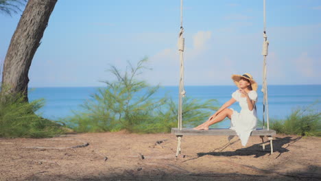 Beautiful-Asian-woman-gently-sitting-on-the-big-wooden-bench-swing-wearing-summer-sundress-and-hat-on-blue-sea-background-in-Thailand,-slow-motion