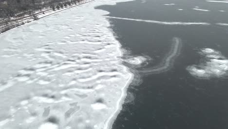 Aerial-view-of-water-partially-frozen,-ice-formations-on-the-shore-with-snow-covering