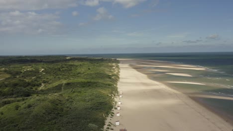 Aerial-shot-moving-slowly-downwards-toward-a-beautiful-beach,-flanked-by-green-dunes-and-a-sea-at-low-tide