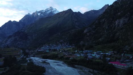 Eine-Drohnenaufnahme-Eines-Kleinen-Malerischen-Abgelegenen-Dorfes-Am-Ufer-Des-Flusses-Baspa-Mit-Den-Schneegipfeln-Des-Indischen-Himalaya-Im-Hintergrund