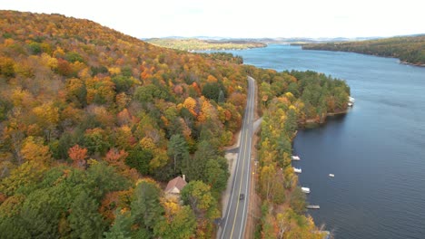 Aerial-View-of-Dark-Vehicle-on-Road-in-Colorful-Autumn-Landscape,-Vivid-Forest-by-Lake-Sunapee,-New-Hampshire-USA,-Tilt-Up-Tracking-Drone-Shot