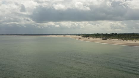 Long-aerial-shot-of-a-coastline-with-a-beach-and-green-dunes-in-Zeeland,-the-Netherlands,-on-a-cloudy-day