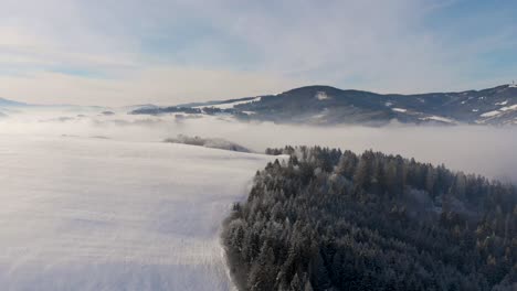 Forward-aerial-drone-over-snowed-in-hill-and-forest-with-mountain-range