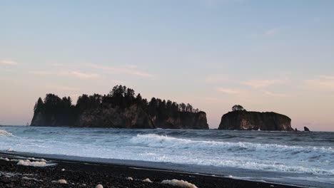 Gorgeous-tilting-up-shot-the-pacific-coastline-with-waves-leaving-behind-sea-foam,-a-beautiful-small-cliff-island,-and-a-colorful-cloudy-sky-during-sunset-at-the-famous-Ruby-Beach-in-Forks,-Washington