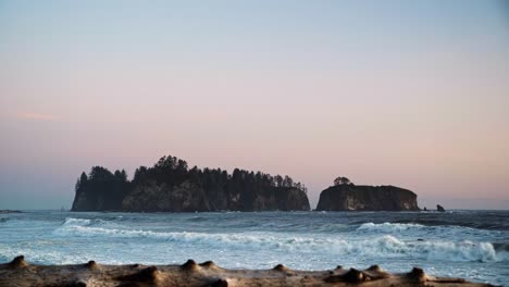 Gorgeous-landscape-beach-shot-of-driftwood-with-waves-crashing-in-the-background,-a-beautiful-small-cliff-island,-and-a-colorful-cloudy-sky-during-sunset-at-the-famous-Ruby-Beach-in-Forks,-Washington