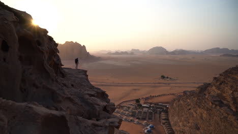 Lonely-man-on-rocky-hill,-viewpoint-of-vastness-of-Wadi-Rum-desert-and-bedouin-camp,-Jordan,-wide-view